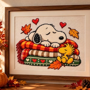 May include: A framed cross-stitch artwork featuring a white dog with black ears sleeping on a stack of colorful blankets, with a small yellow bird sitting beside it. The design includes red hearts and orange autumn leaves on a white grid background.