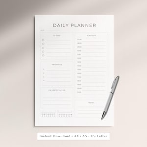 Op de afbeelding: Een witte dagplanner met secties voor taken, schema, prioriteiten en notities. Een zilveren pen ligt aan de rechterkant. De tekst "DAILY PLANNER" staat bovenaan. De onderkant van de planner luidt "Instant Download • A4 • A5 • US Letter".