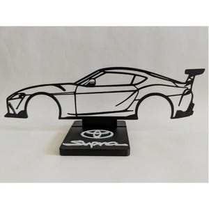 Puede incluir: Una silueta de metal negro de un coche deportivo Toyota Supra montada sobre una base rectangular negra. La base presenta el logotipo blanco de Toyota y la palabra Supra en letra cursiva blanca. El contorno incluye un alerón trasero.
