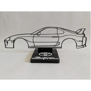 Może przedstawiać: Czarna rzeźba z drutu przedstawiająca sylwetkę samochodu sportowego Toyota Supra, umieszczona na prostokątnej czarnej podstawie. Na górnej powierzchni podstawy znajduje się białe logo Toyoty oraz napis Supra wykonany białą czcionką.