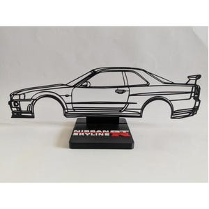 Può includere: Una scultura in filo metallico nero che raffigura il profilo di un'auto Nissan Skyline GT-R, montata su una base rettangolare nera. La base presenta la scritta bianca NISSAN SKYLINE con un logo R rosso. La scultura mostra la sagoma laterale.