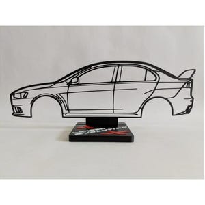 Può includere: Una scultura in filo metallico nero di una berlina Mitsubishi Lancer Evolution X vista di profilo. Il contorno dell'auto è montato su una base rettangolare nera con il testo rosso e bianco Lancer Evolution X stampato sulla superficie.