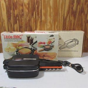 May include: A black and silver Little Mac 60 Second Burger Machine with its original box. The box features images of burgers and other food items. The machine has a black handle and a power cord.