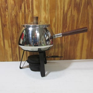 May include: A vintage stainless steel fondue pot with a wooden handle and lid. The pot sits on a black metal stand with a fuel burner. The background is a wooden wall.