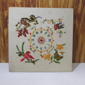 May include: Embroidered clock face with Roman numerals surrounded by floral and bird motifs. The clock face is white with black Roman numerals and a red center. The floral motifs are in shades of yellow, red, and green. The birds are red and brown.