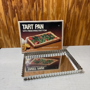 May include: A rectangular tart pan with a removable bottom, made of shiny tinplate. The box behind it reads "TART PAN WITH REMOVABLE BOTTOM". The pan has a fluted edge and is designed for baking. The box also says "PROFESSIONAL QUALITY TINPLATE".