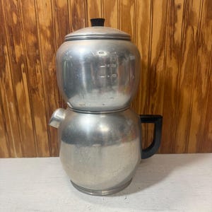 May include: A vintage aluminum coffee percolator with a black handle and lid. The coffee pot has a two-part design with a spout and measurement markings on the side. The percolator sits on a white surface against a wooden background.