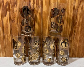 May include: Set of six vintage clear drinking glasses. Each glass is decorated with gold and black coin designs. The glasses are cylindrical and appear to be made of glass. The glasses are arranged in a stack on a wooden surface.