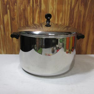 May include: A stainless steel cooking pot with a lid. The pot has a shiny, reflective surface and black handles. The lid has a black knob. The pot is sitting on a white surface against a wooden background.