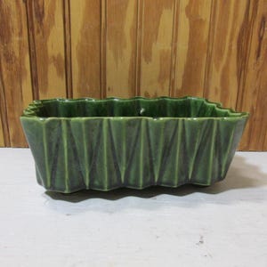 May include: A rectangular, dark green ceramic planter with a textured, geometric design. The planter has a wavy top edge and is suitable for indoor or outdoor use. The planter is approximately 10 inches long.