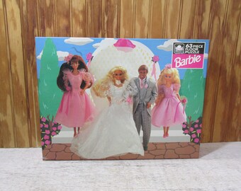 May include: A 63-piece floor puzzle featuring Barbie dolls in a wedding scene. The bride is wearing a white dress, the groom is wearing a gray suit, and two other dolls are wearing pink dresses. The box is blue and pink with the text "63 piece floor puzzle Barbie" on it.