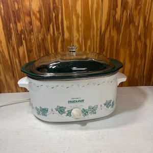 May include: A white Rival Crock-Pot stoneware slow cooker with a green ivy leaf pattern, a clear glass lid, and a white control knob. The front features the Rival Crock-Pot logo and settings for off, low, and high heat, with white side handles.