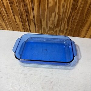 May include: A rectangular cobalt blue glass baking dish with integrated side handles. The center of the dish features the embossed text Pyrex, indicating the brand. The glass is transparent and has a smooth, polished finish throughout.