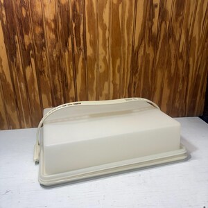 May include: A rectangular, off-white plastic cake carrier with a flat base and a removable lid. The lid features a curved, integrated handle that arches over the top, designed for secure transport of baked goods in a kitchen setting.
