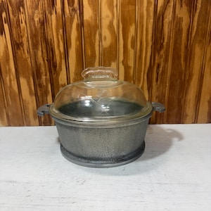 May include: A round, gray, textured metal pot with two side handles and a clear, domed glass lid. The lid features a small, etched floral design and a central handle. The pot has a wide, flat base and a consistent, speckled metallic finish.