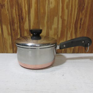 May include: A small, stainless steel saucepan with a copper-colored base and a lid. The pan has a black handle and a black knob on the lid. The pan is designed for cooking and is likely used in a kitchen setting.