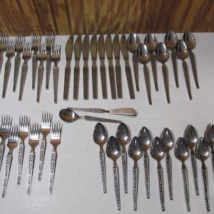 May include: A collection of vintage silverware, including forks, knives, and spoons, arranged on a white surface. The silverware has a decorative pattern on the handles. The spoons have a rounded bowl shape.