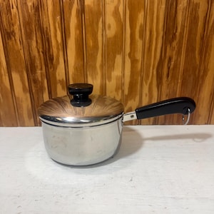 May include: A small, stainless steel saucepan with a lid and a black handle. The lid has a black knob. The pan is sitting on a white surface with a wooden background.
