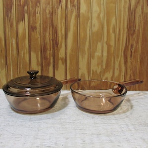May include: Two pieces of amber-colored glass cookware. One piece is a round saucepan with a matching glass lid and a side handle. The second piece is an open glass skillet with a side handle. Both items feature a smooth, translucent finish.