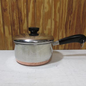 May include: A stainless steel saucepan with a copper-colored base and a lid. The pan has a black handle and a black knob on the lid. The pan is sitting on a white surface in front of a wooden background.
