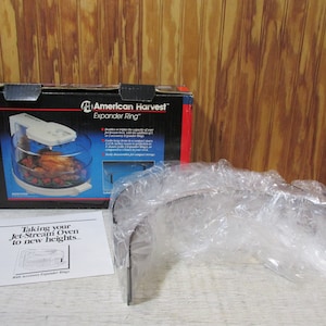 May include: A white and black box with the words "American Harvest Expander Ring" on it. The box shows a clear glass oven with a roasted chicken inside. The box is sitting on a wooden surface with a clear plastic bag next to it. The bag contains a metal ring.