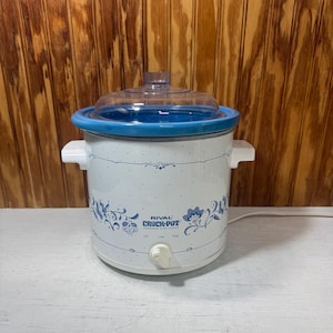 May include: White Rival Crock-Pot slow cooker with blue floral design and a clear glass lid. The slow cooker has a blue rim and white handles. The control knob has settings for Off, Low, and High.