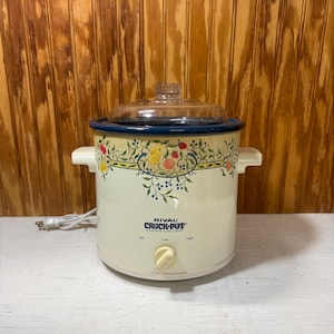 May include: A cream-colored Rival Crock-Pot slow cooker with a clear glass lid and a blue rim. The exterior features a decorative fruit and vine pattern. It has a front-facing control knob with Off, Low, and High settings and a white power cord.