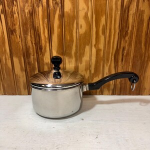 May include: A stainless steel saucepan with a black handle and lid. The lid has a black knob. The saucepan is sitting on a white surface with a wooden background. The handle has a ring at the end.