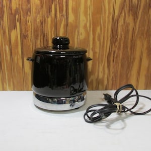 May include: A black electric fondue pot with a lid and side handles. The pot sits on a chrome base and has a black power cord. The background is a wooden wall.