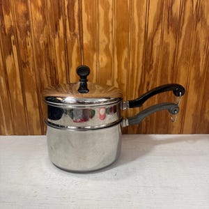 May include: A stainless steel double boiler with a lid and two black handles. The pot has a shiny, reflective surface and sits on a white surface against a wooden background. The lid has a black knob.