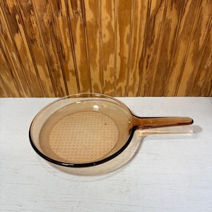 May include: A round, amber-colored glass skillet with a long, integrated handle. The base of the pan features a subtle, raised grid pattern texture, while the sides and handle are smooth and translucent, allowing light to pass through the glass.