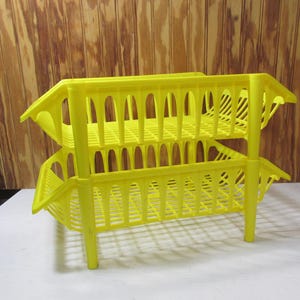 May include: A bright yellow, two-tiered storage rack. The rack is made of plastic and has a rectangular shape with a series of vertical slots. The rack is supported by four legs. The background is a wooden wall.