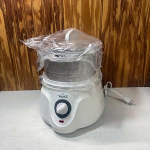 May include: A white Rival brand electric food steamer with a clear plastic lid and container, partially wrapped in protective plastic film. The base features a dark gray circular control dial with a white knob and a small red power indicator light.