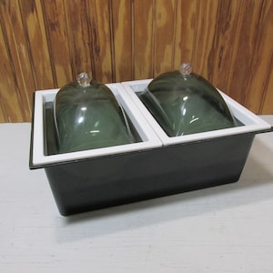 May include: A dark gray serving dish with two compartments, each holding a green glass cloche. The cloches have clear glass knobs. The dish has a white interior and a black exterior.