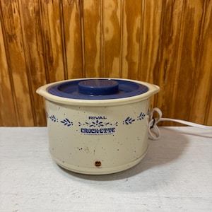 May include: A cream-colored Rival Crock-Ette stoneware slow cooker with a blue lid and blue floral pattern. The exterior features a speckled finish, a small orange indicator light, and a white power cord extending from the side of the unit.
