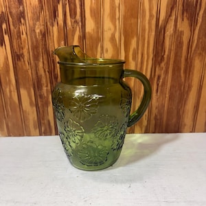 May include: A translucent olive green glass pitcher featuring a repeating embossed daisy flower pattern across its exterior surface. The pitcher includes a curved handle on the right side and a wide pouring spout at the top rim of the vessel.