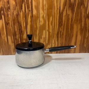 May include: A silver-toned metal saucepan with a smooth, rounded body and a long, black handle. The pot features a matching black lid with a small, tapered black knob on top. The handle includes a silver-colored metal end cap for hanging.