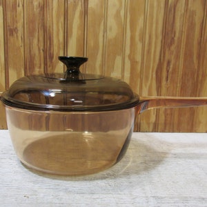 May include: A transparent amber-colored saucepan with a matching lid and a black handle. The saucepan has a long handle and a rounded shape. The lid has a black knob. The background is a light brown wooden surface.