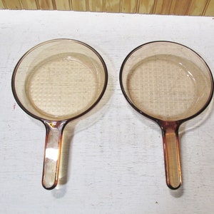 May include: Two round, amber-colored glass skillets with long, integrated handles. Each skillet features a smooth, circular basin with a subtle grid-like texture visible on the bottom surface, and the glass is translucent with a warm brown tint.