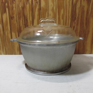 May include: A vintage, silver-colored cooking pot with a clear glass lid. The lid has a decorative handle and etched leaf design. The pot has two side handles and sits on a round base. The background is a wooden wall.
