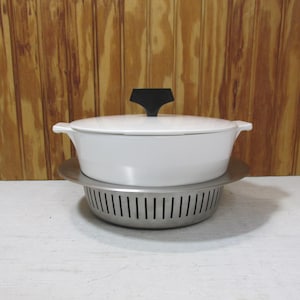May include: A white enameled cast iron casserole dish with a black handle on the lid. The dish sits on a stainless steel trivet with a perforated design. The background is a wooden wall.