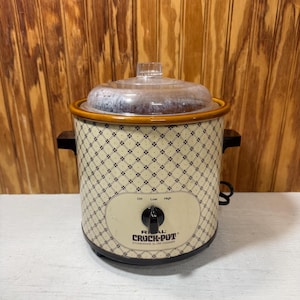 May include: Cream-colored Rival Crock-Pot stoneware slow cooker with a clear glass lid. The slow cooker has a black and cream diamond pattern, black handles, and a control knob with Off, Low, and High settings.