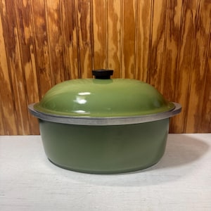 May include: A green enamel-coated cooking pot with a lid. The pot has a silver rim and a black knob on top of the lid. The pot is sitting on a white surface with a wooden background.