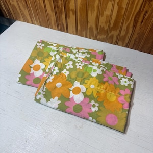 May include: Two folded pillowcases with a floral pattern. The pattern features large orange, pink, and white flowers on a green background. The pillowcases are made of a soft fabric and are likely vintage.