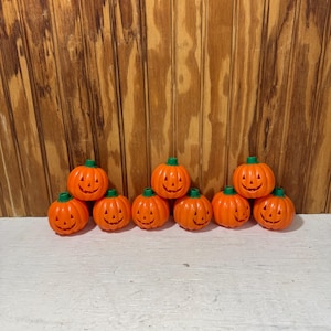May include: A collection of eight small, bright orange plastic jack-o'-lanterns arranged in a row. Each pumpkin features a carved face with triangular eyes, a triangular nose, and a jagged mouth, topped with a small, solid green stem.