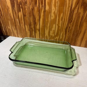 May include: A rectangular, light green glass baking dish with a black trim. The dish has handles on each end and is empty. The dish is sitting on a white surface with a wooden background.