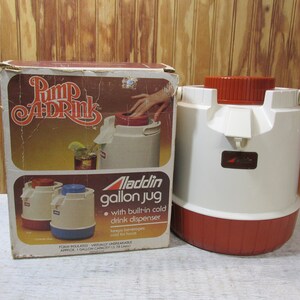 May include: A white and red Aladdin gallon jug with a built-in cold drink dispenser. The jug is on a wooden surface and has the text "Pump A Drink" on the box. The jug is foam insulated and virtually unbreakable.