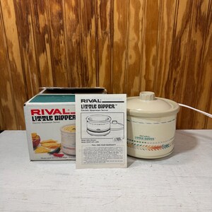 May include: A cream-colored Rival Little Dipper electric stoneware server with a matching lid, accompanied by its original cardboard box and a printed instruction manual. The server features a decorative band with geometric patterns in muted colors.