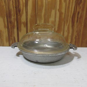 May include: A vintage, round, metal casserole dish with a clear glass lid. The dish has two handles and a clear glass lid with a handle. The dish is silver in color and sits on a white surface.