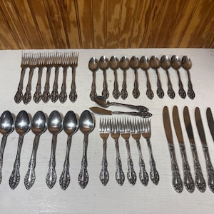 May include: A collection of vintage silverware, including forks, spoons, knives, and butter knives, arranged on a white surface. The silverware has ornate, floral-like detailing on the handles, and a shiny, silver finish.